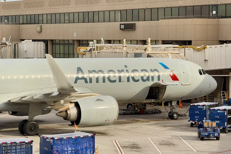 An American Airlines aircraft parked at the gate at an airport, with ground service vehicles and baggage carts positioned nearby. The plane’s cargo door is open as it is being loaded or serviced before departure.