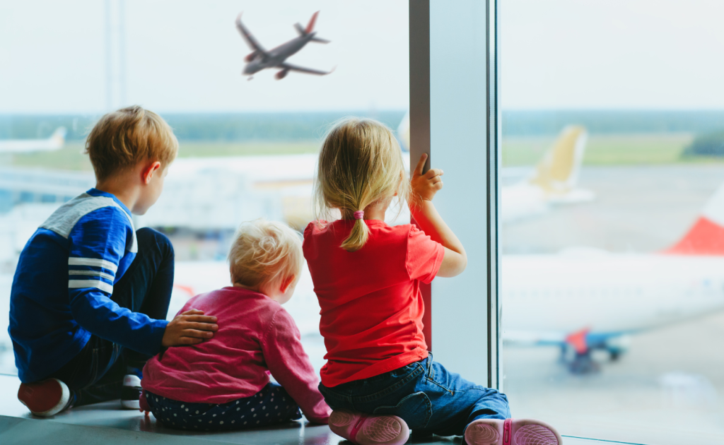 Three children sitting by an airport window watching airplanes take off on the runway possibly boarding early