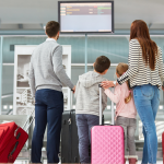 Family with children boarding early without paying extra at an airport, looking at a flight information display while holding suitcases.