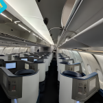 Empty first-class airplane cabin with private lie-flat pods and screens. Bold overlay: "FIRST CLASS UPGRADES!"