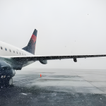 Delta airplane on snowy, wet tarmac in foggy weather, with ice on wing and engine, orange cones marking area illustrating weather related flight delays or cancellations.
