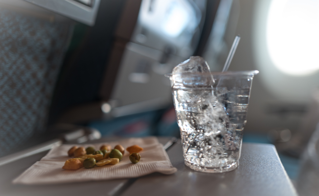 Plastic cup of water with ice on an airplane tray table beside snacks.