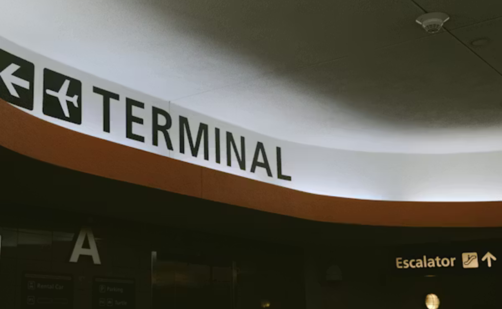 Overhead illuminated sign in an airport directing to "Terminal A" with airplane icon and arrow left, escalator sign visible below with TSA fees