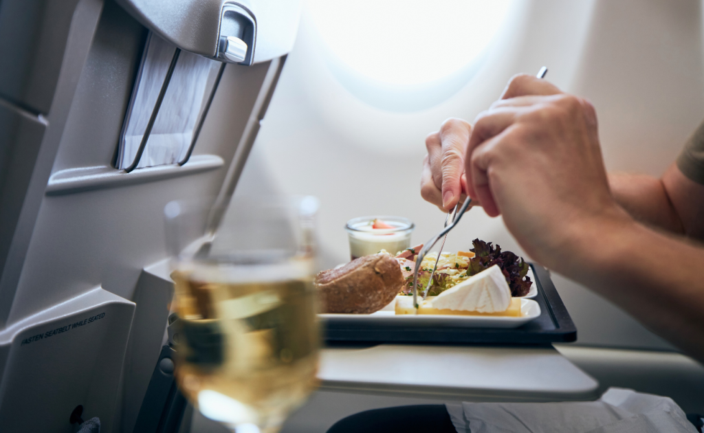 Passenger eating an in-flight meal with a glass of alcohol on an airplane tray table.