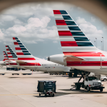View through an airplane window of several American Airlines planes with iconic striped tails parked on the tarmac, ground crew and equipment visible under partly cloudy skies.