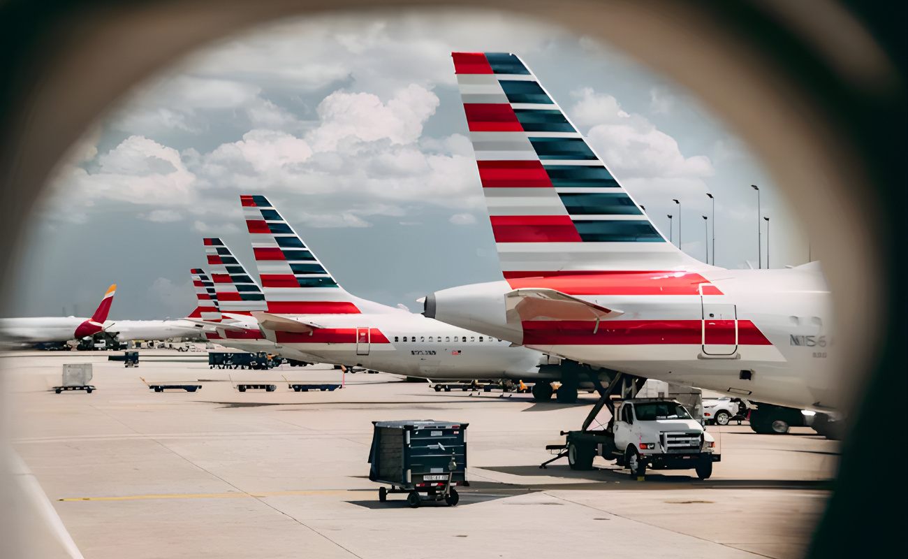 View through an airplane window of several American Airlines planes with iconic striped tails parked on the tarmac, ground crew and equipment visible under partly cloudy skies.