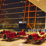 Nearly empty airport gate E19 at night with rows of red seats, one lone passenger sleeping with suitcase nearby due to his delayed flights.