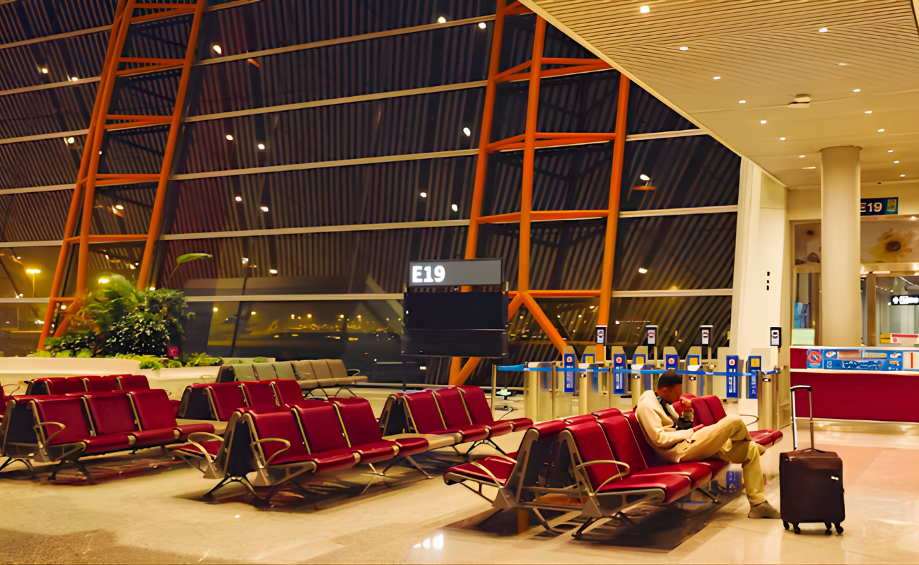 Nearly empty airport gate E19 at night with rows of red seats, one lone passenger sleeping with suitcase nearby due to his delayed flights.