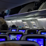 Packed airplane cabin full of seated passengers, flight attendant standing in aisle preparing for takeoff.