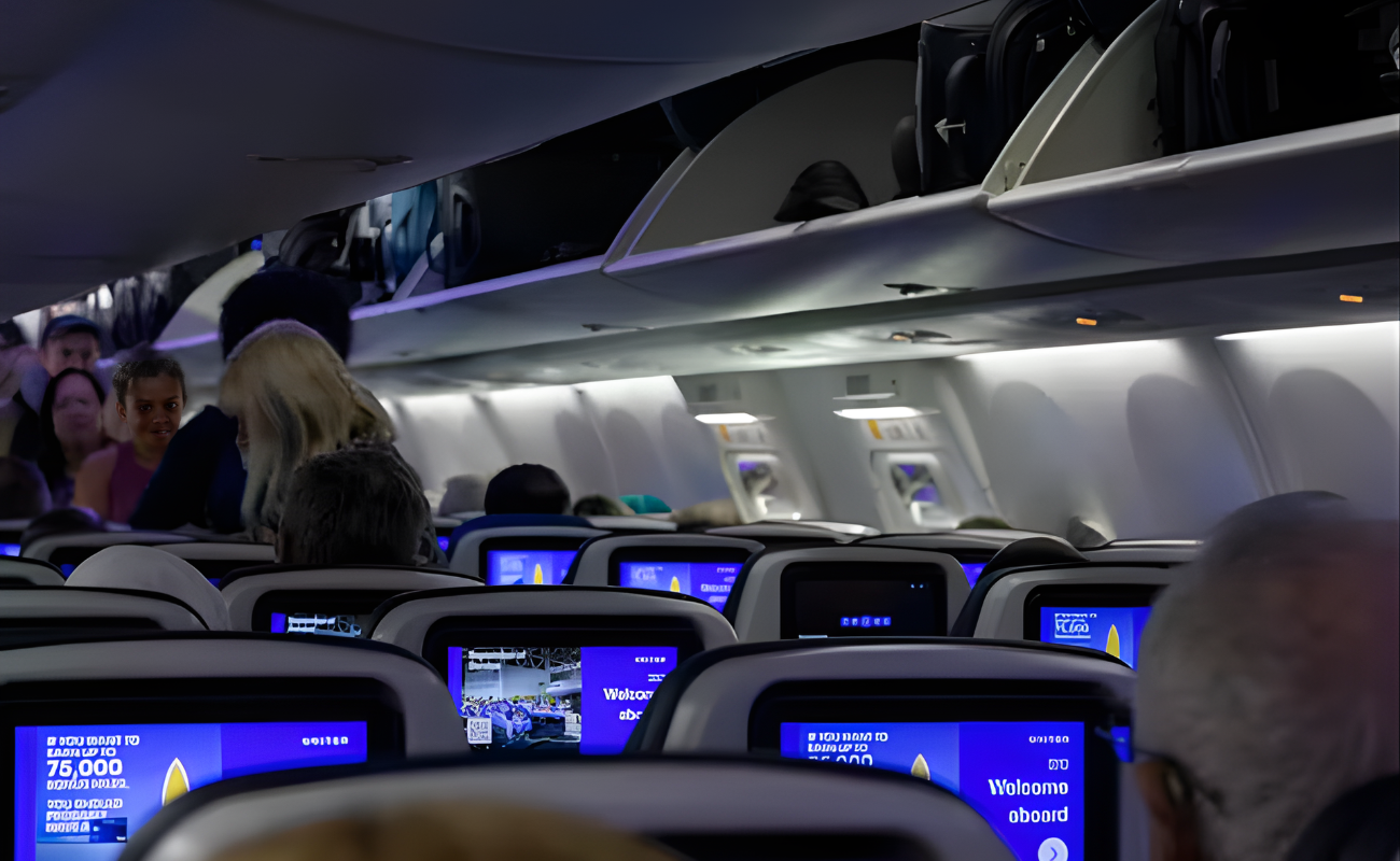 Packed airplane cabin full of seated passengers, flight attendant standing in aisle preparing for takeoff.