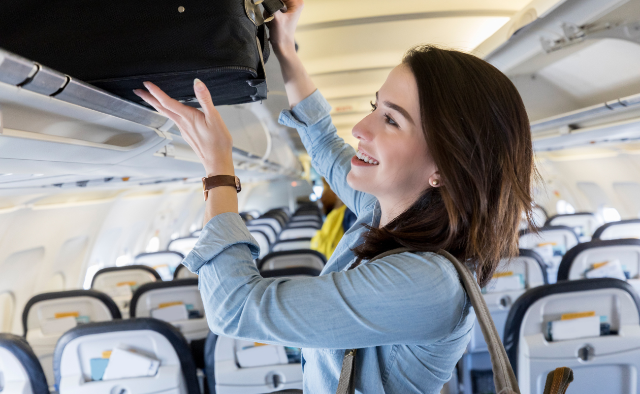 Woman placing a carry-on bag into an airplane overhead bin during boarding.