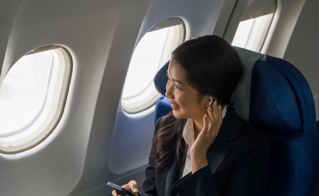 Passenger wearing wireless bluetooth earbuds and using a smartphone while seated by an airplane window.