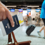Hand holding U.S. passport with boarding pass tucked in, luggage handle in grip, blurred airport terminal background.