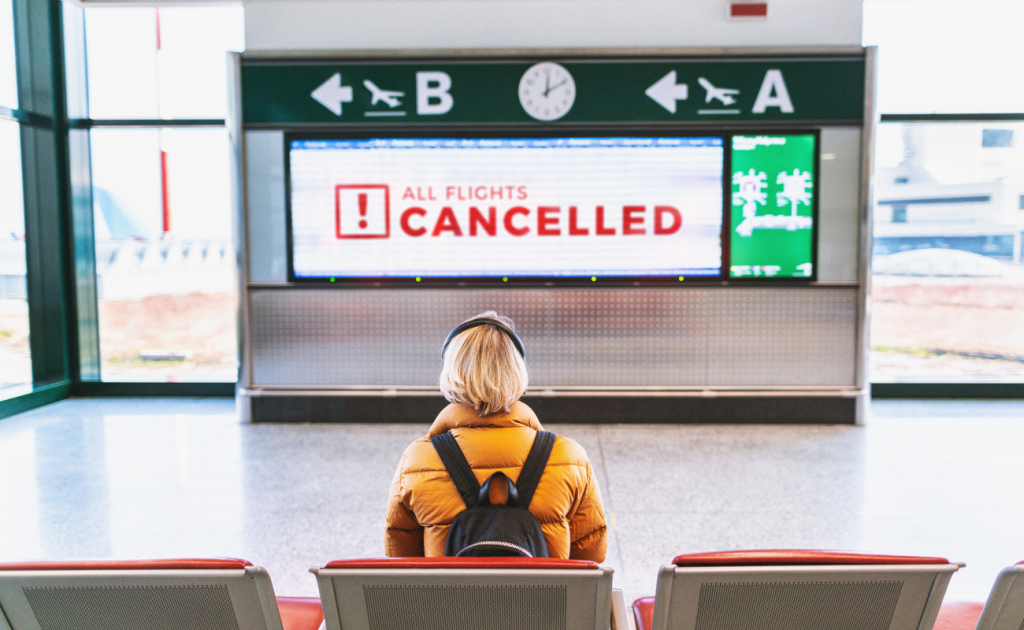 Traveler sitting in airport terminal facing sign that says all flights cancelled.
