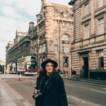 Woman wearing black coat and hat holding camera on a European city street lined with historic buildings and buses