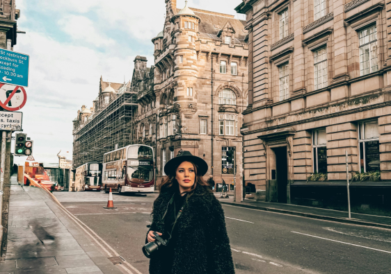 Woman wearing black coat and hat holding camera on a European city street lined with historic buildings and buses