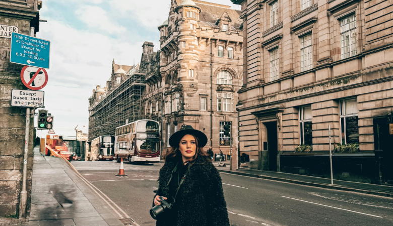 Woman wearing black coat and hat holding camera on a European city street lined with historic buildings and buses