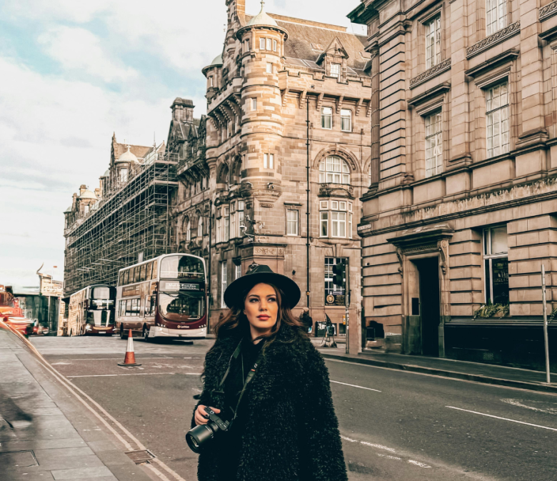 Woman wearing black coat and hat holding camera on a European city street lined with historic buildings and buses