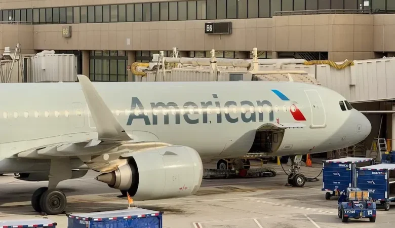 An American Airlines aircraft parked at the gate at an airport, with ground service vehicles and baggage carts positioned nearby. The plane’s cargo door is open as it is being loaded or serviced before departure.